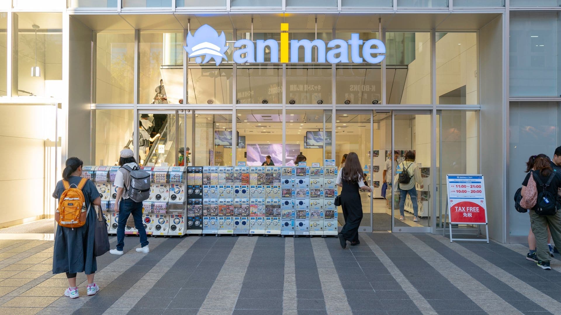 Glass-fronted store with a sign that reads 'animate' and a logo of a blue, stylized flame. Several people are outside, some looking at vending machines, and others entering the store. A red and white sign indicates tax-free shopping.
