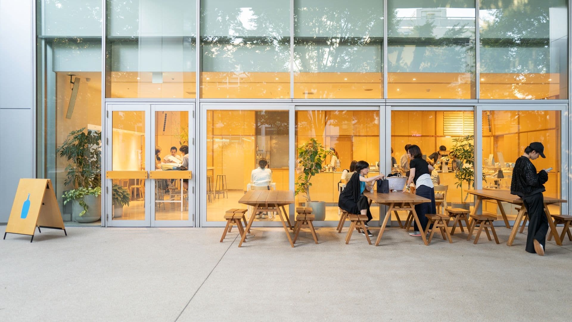 Outdoor patio seating area outside a modern cafe with people sitting at wooden tables and staff attending to customers, large glass windows with warm lighting inside.