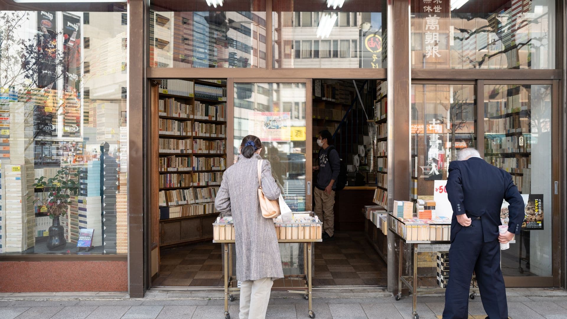People browsing books outside a bookstore with large glass windows, displaying books and a plant inside.
