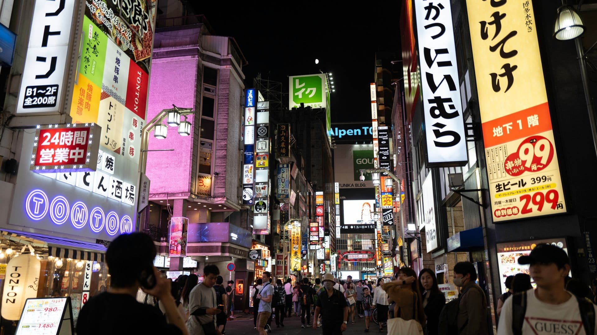 A busy city street at night in Japan with bright neon signs, advertisements, and many pedestrians.