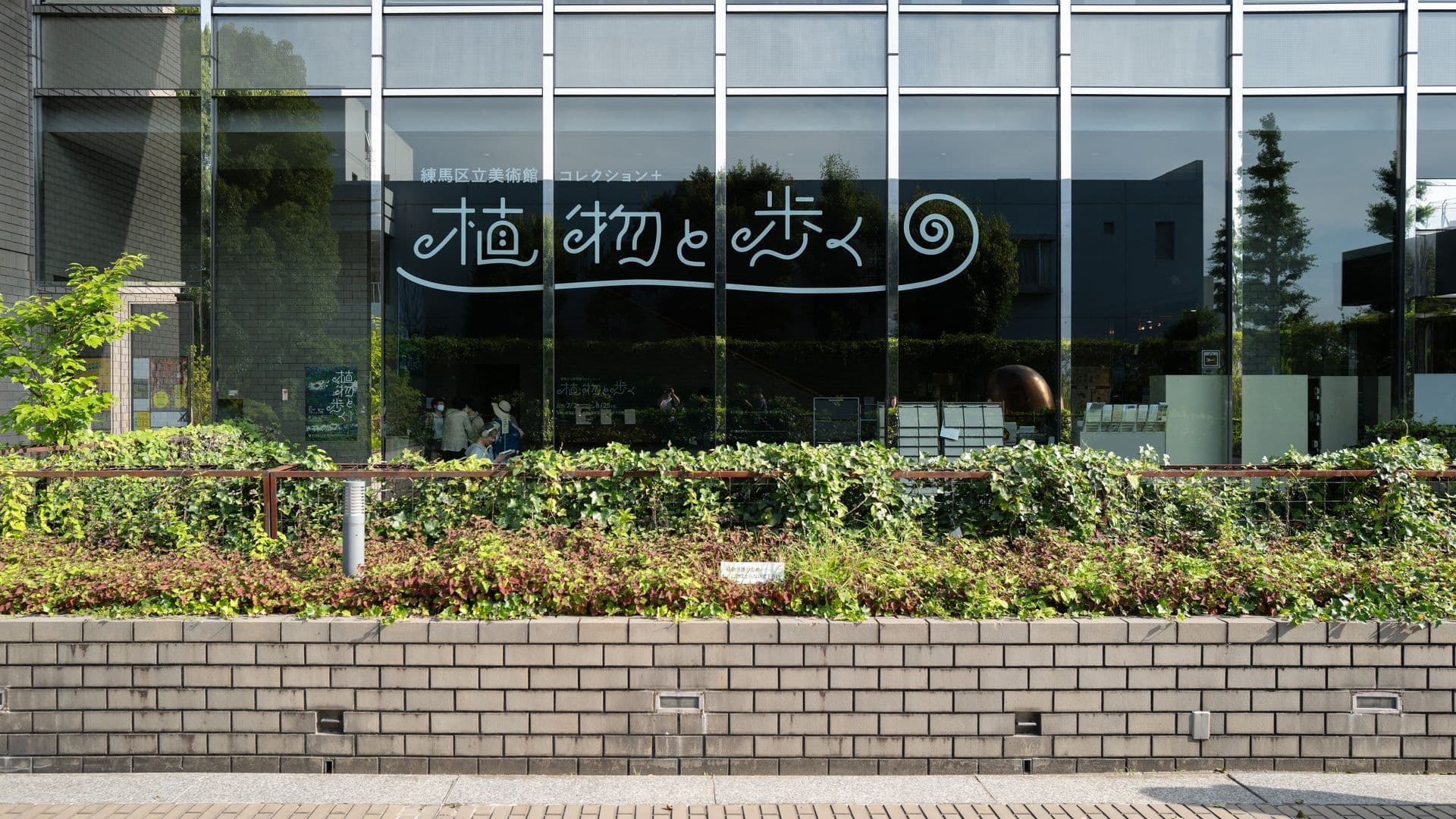 A modern glass building with Japanese characters and a spiral design on the window, reflecting trees and a blue sky, with a garden and brick wall in the foreground.