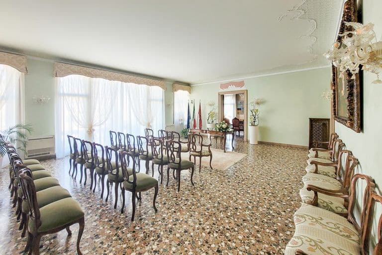 Elegant conference room with chairs arranged around a central table, large windows with sheer curtains, decorative ceiling molding, and ornate lighting fixtures.