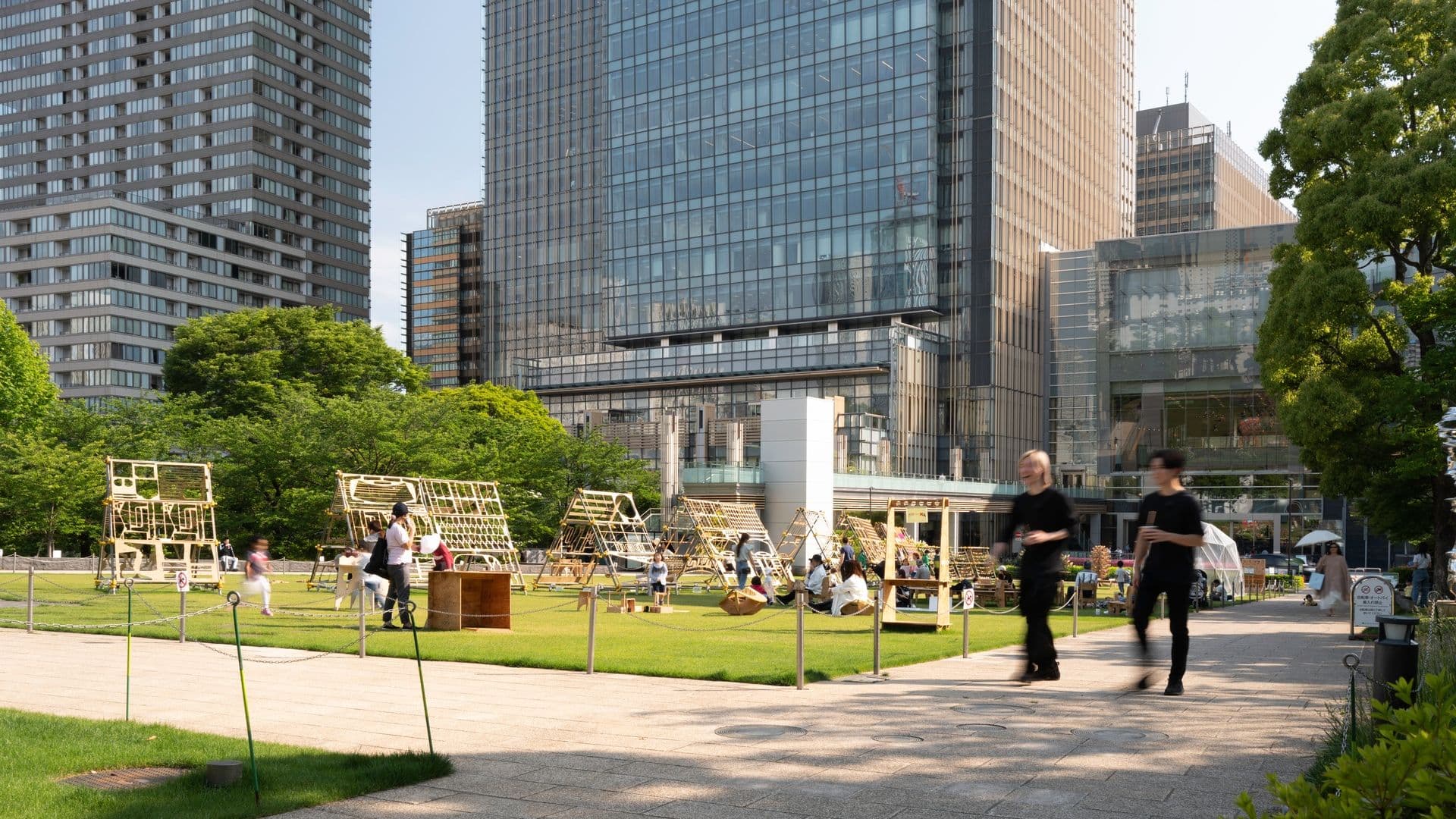 Urban park with children and adults playing and relaxing on grassy areas, modern glass skyscrapers in the background, and people walking on a paved pathway.