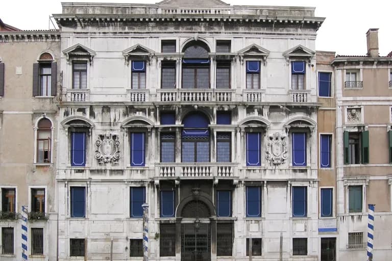 Historical building with decorative windows and balconies, showing signs of dirt and weathering, located along a canal with striped posts in Venice.