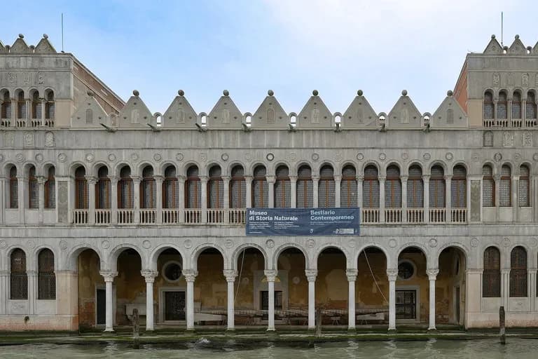 Historic Venetian building with multiple archways and ornate windows, situated along a canal with wooden posts in the water.