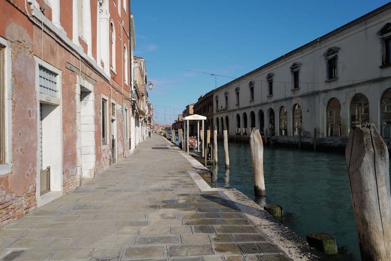 A narrow riverside street with old buildings, some with peeling paint, and a canal with wooden posts in Venice, Italy, under a clear blue sky.