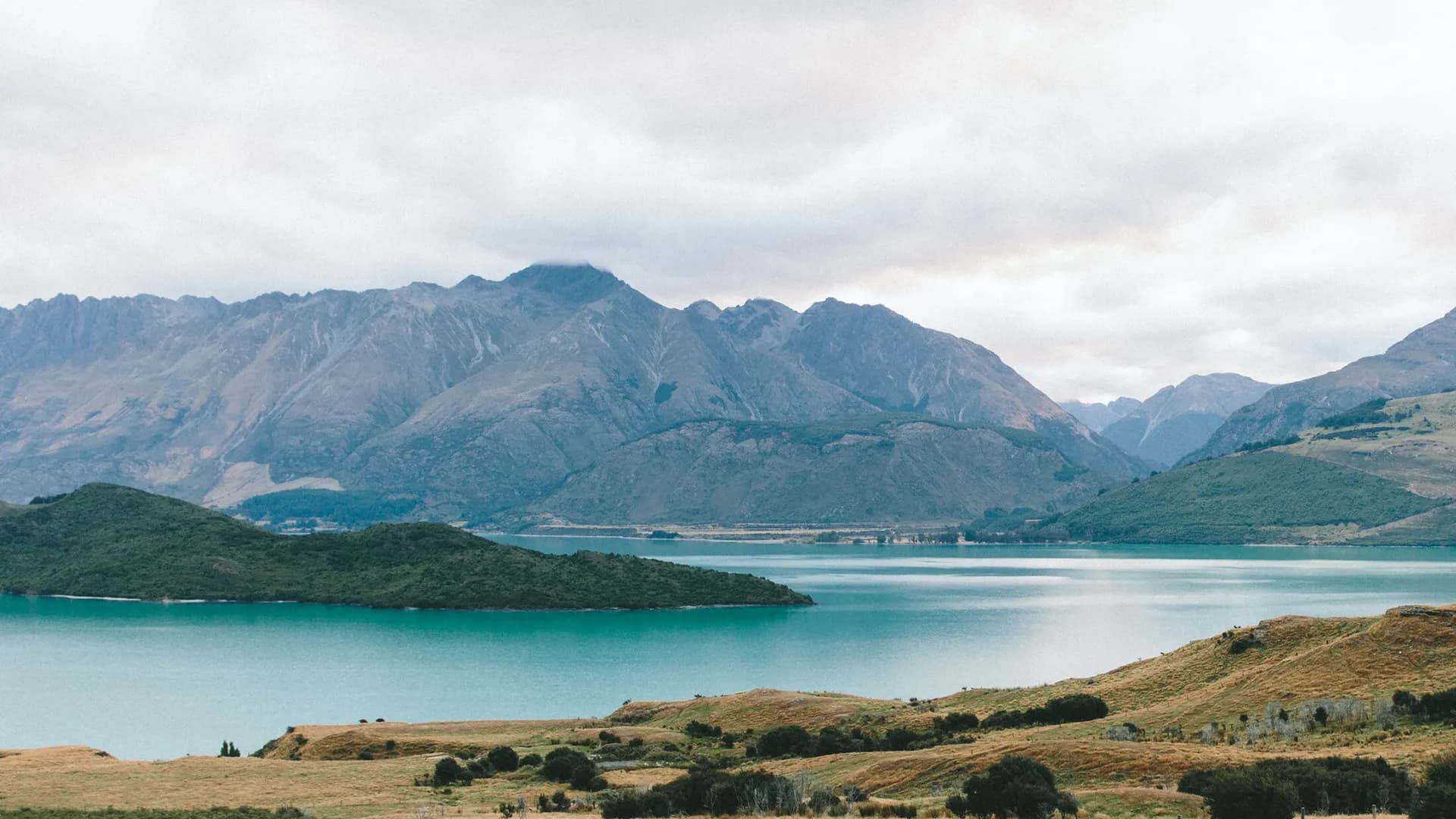 Scenic landscape of a lake with green islands, surrounded by mountains with cloudy skies