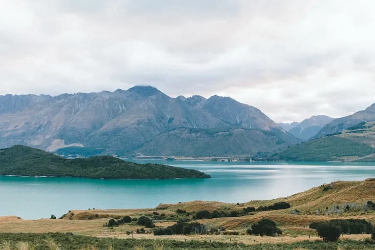 Scenic view of turquoise lake surrounded by mountains and grassy hills under a cloudy sky.
