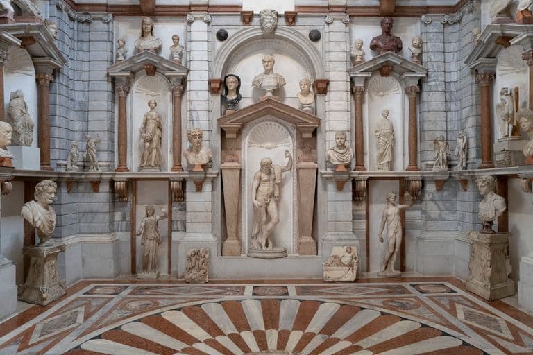 Interior view of a museum hall featuring numerous classical statues and busts displayed on shelves and pedestals against a marble wall.