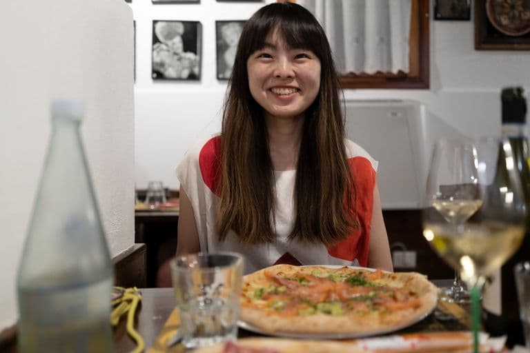 Woman smiling at a restaurant table with a pizza and a glass of white wine in front of her.