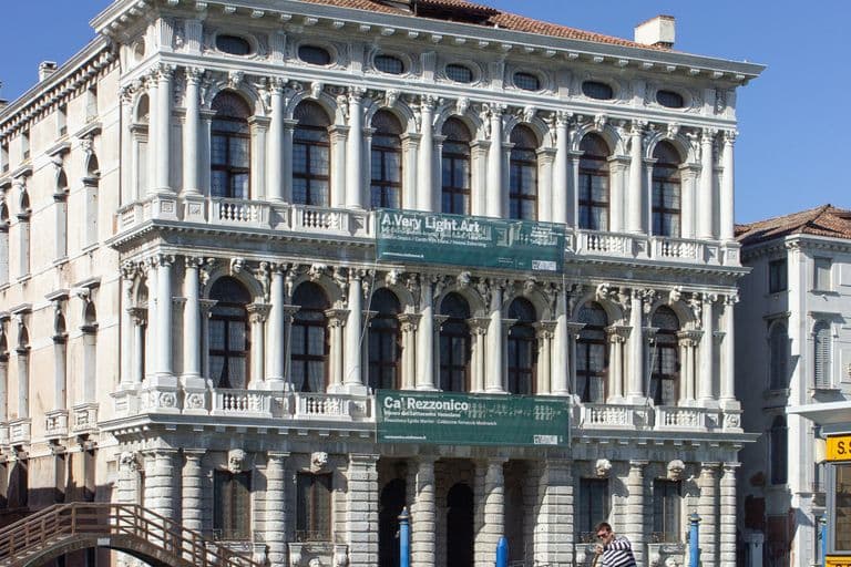 A historic white building with ornate architecture, situated along a canal, with a gondola and gondolier in Venice, Italy.