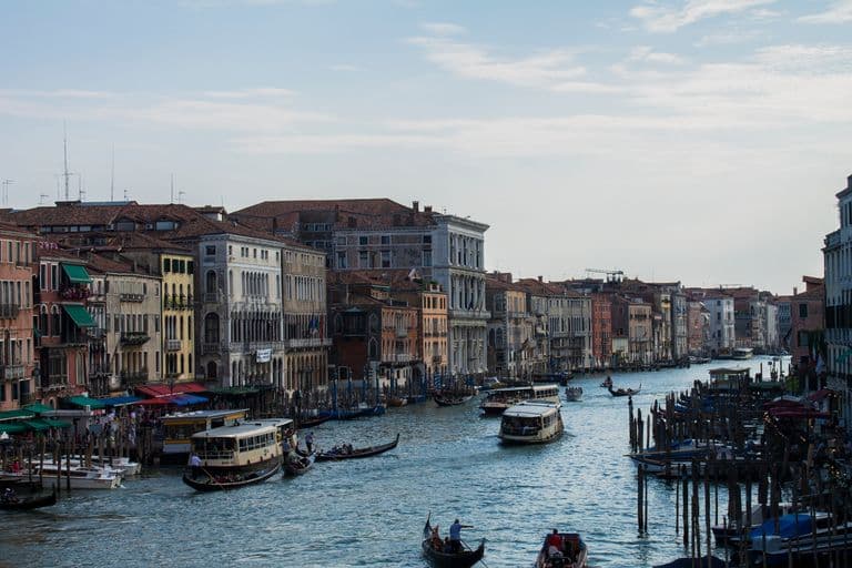 A view of the Grand Canal in Venice, Italy with historic buildings on either side, boats including gondolas and water taxis on the water, under a partly cloudy sky.