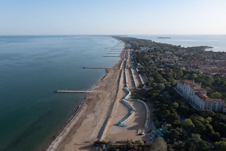 Aerial view of a coastline under construction with beach, shoreline, and a row of buildings and green trees along the land. Promenade and beach chairs are visible.