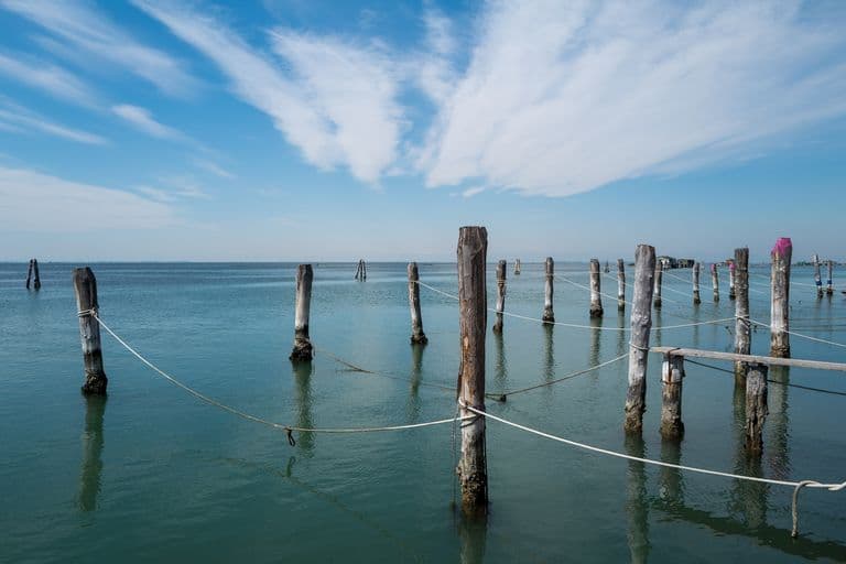 A calm body of water with weathered wooden posts connected by ropes, some with pink paint, under a partly cloudy blue sky.