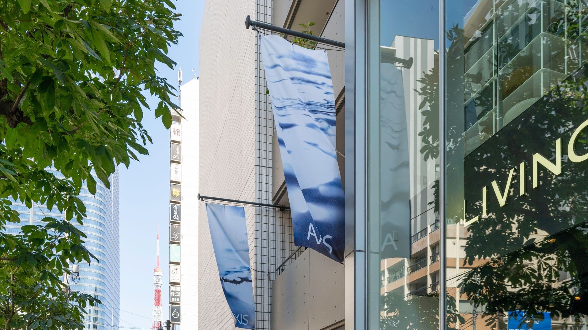 Street view of a modern building with glass windows, displaying banners with blue and white designs, and surrounding trees and tall buildings in an urban setting.