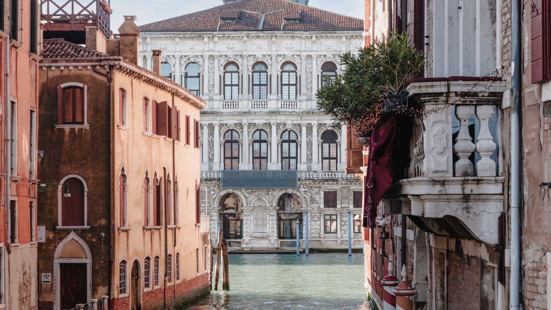 View of narrow canal between historic buildings in Venice, Italy, with the Grand Canal and ornate white building in the background.