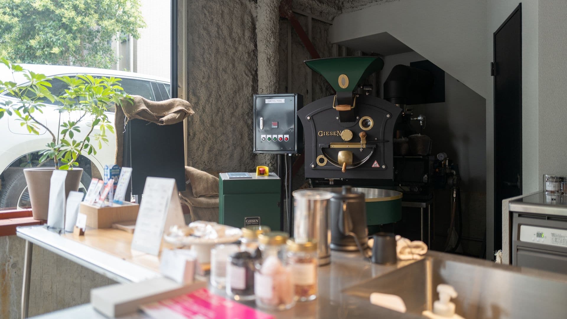 Coffee roasting machine in a cozy coffee shop with a large window, a potted plant, and various coffee equipment on the counter.