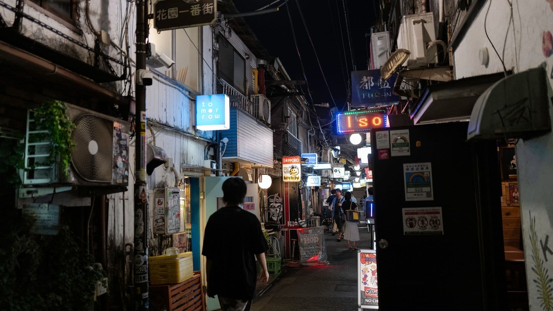 Nighttime scene of a narrow street with illuminated signs and people walking, featuring small shops and restaurants with various neon and LED lights in an urban setting.