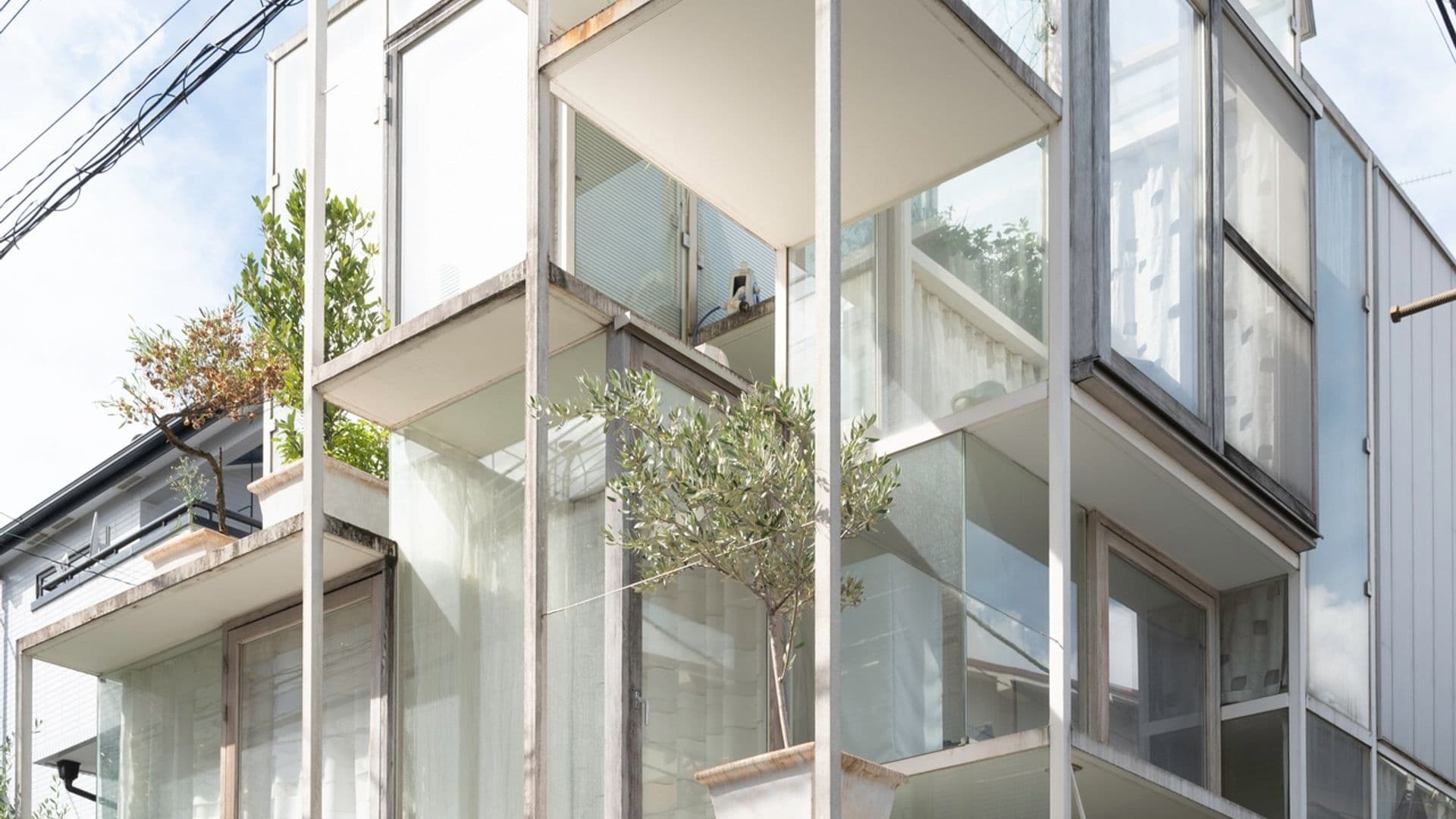Modern multi-story building with glass balconies and potted plants, with a vintage white car and bicycles parked underneath, and power lines overhead.