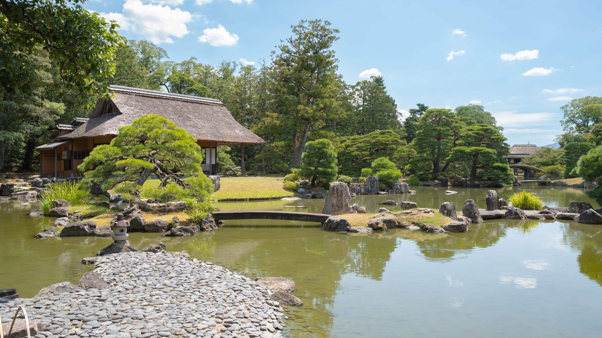 A traditional Japanese garden with a pond, stone pathway, lush green trees, and a thatched-roof house in the background under a partly cloudy sky.