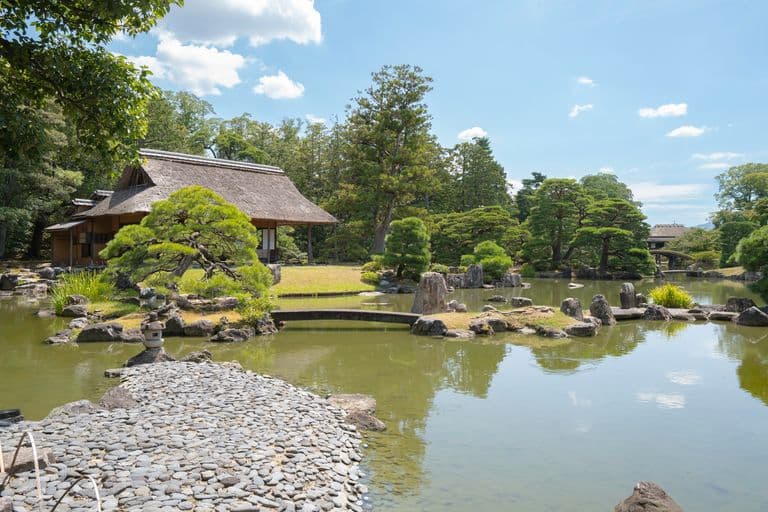 Traditional Japanese garden with pond, stone pathway, pine trees, and a thatched-roof house under a blue sky.