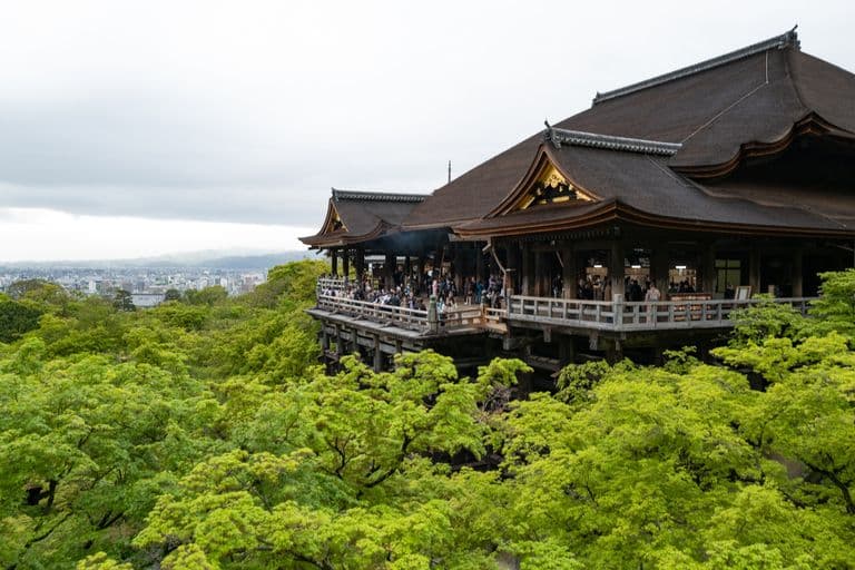Traditional Japanese wooden temple on a hill with lush green trees and a city skyline in the background under an overcast sky.