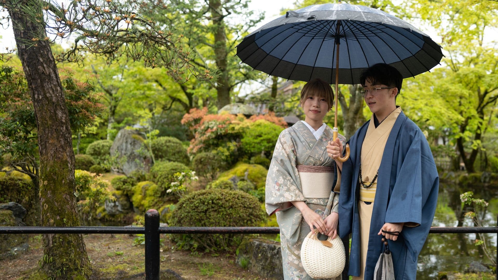 A young woman and a young man standing under a black umbrella in a lush, green Japanese garden. They are wearing traditional Japanese kimonos, and the woman is holding a woven handbag while the man holds a black and white striped bag. The garden features trees, shrubs, and rocks, with a small pond visible in the background.