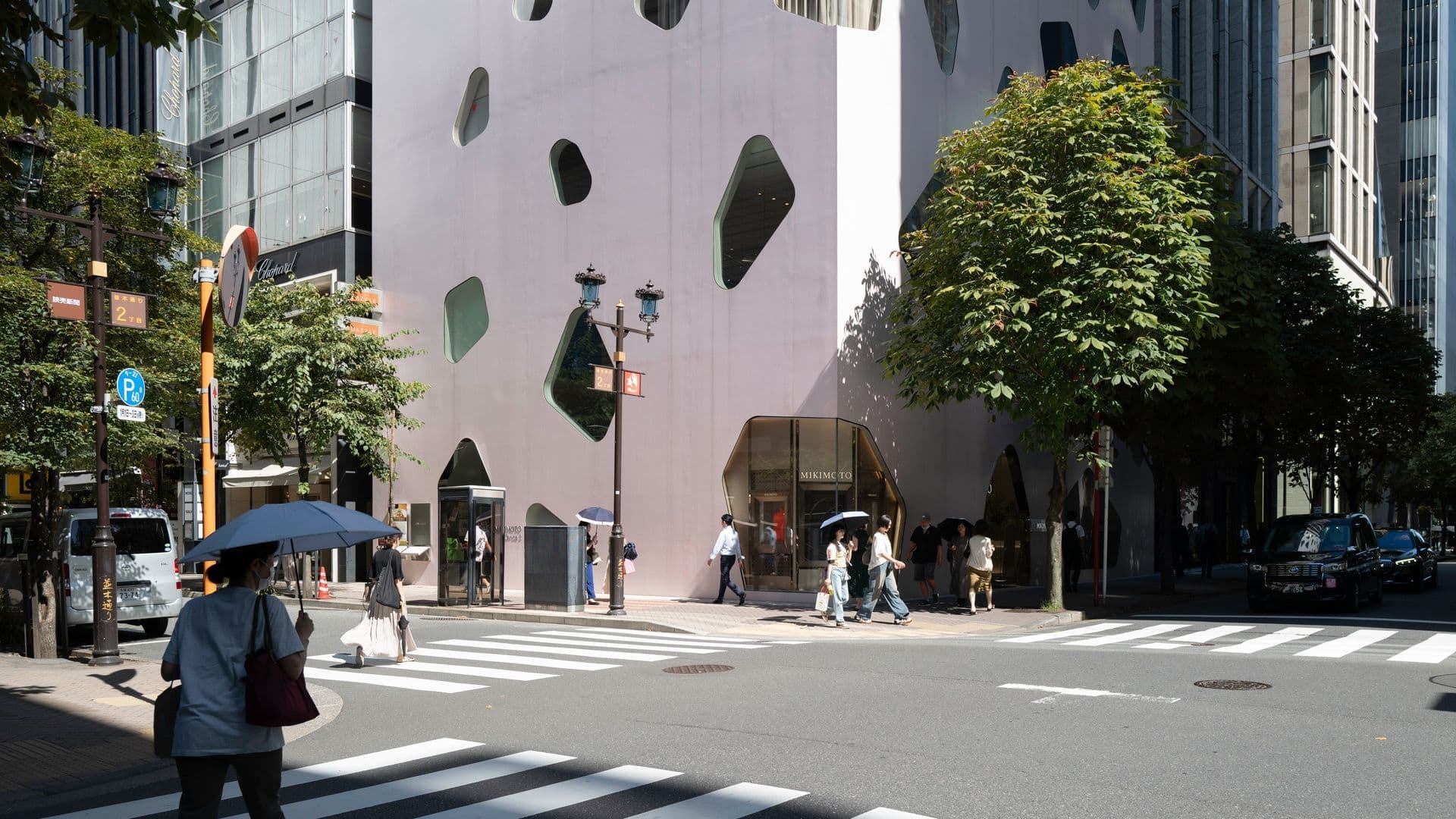 People walking across a crosswalk in front of a modern building with irregularly shaped windows and trees lining the sidewalk.