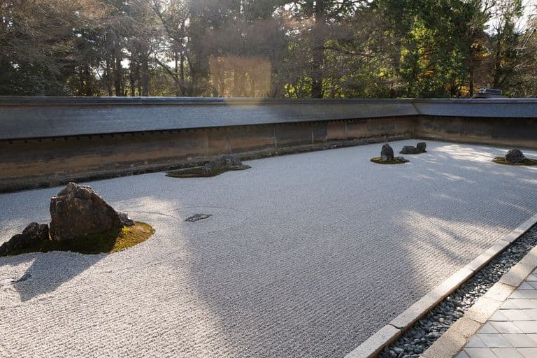 Japanese Zen garden with white gravel and several large rocks, bordered by a low stone wall and surrounded by trees.