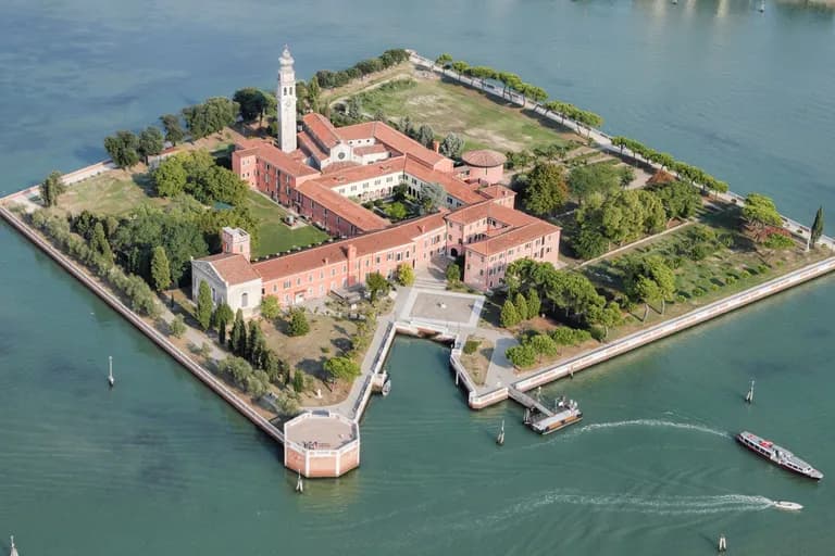 Aerial view of a square-shaped island in Venice with historical buildings, a tall clock tower, courtyards, and surrounding gardens. Boats are traveling in the surrounding water.
