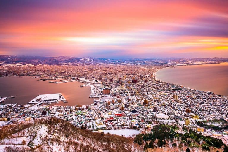 Aerial view of a snow-covered city with a harbor and mountains in the background during a colorful sunset sky.