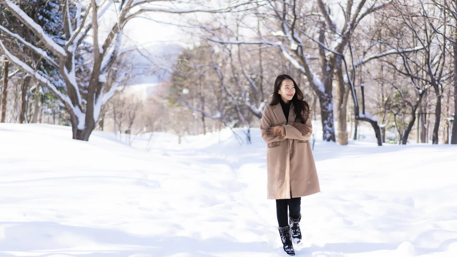 A woman in a tan coat and black boots walking through a snowy park with trees covered in snow, smiling and looking to the side.